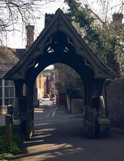 A detailed view of an ornate Gothic-style wooden archway serving as an entrance to a garden or courtyard, featuring intricate carving and decorative elements along the roofline. Beyond the arch, a nar