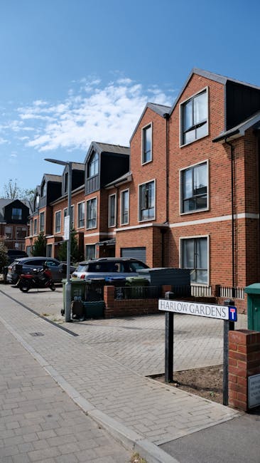 A modern residential street in Harrow Weald featuring multi-story brick buildings with black accents and large windows, under a bright blue sky with scattered white clouds. In the foreground, a paved